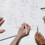 Dua orang berkolaborasi di meja kerja dengan gambar teknis dan prototype produk